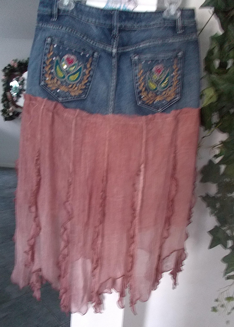 Op de afbeelding: Een spijkerbroekrok met een vervaagde blauwe wassing en een roze, vloeiende stoffen onderkant. Het denimgedeelte heeft geborduurde bloemmotieven op de achterzakken. De rok heeft een uniek, gelaagd ontwerp dat casual denim combineert met een zacht, vloeiend materiaal.