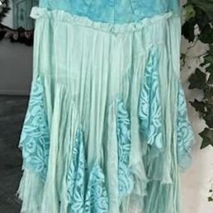 Puede incluir: Falda de mezclilla y encaje en tonos azules y turquesas. La falda presenta una cintura de mezclilla y un dise&ntilde;o fluido y en capas con detalles de encaje. La falda est&aacute; colgada en una percha.