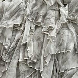May include: Close-up of a layered, flowing garment with a marbled gray and white pattern. The fabric is sheer and ruffled, creating a textured effect. The top edge features a decorative embellishment with silver and pearl-like beads.