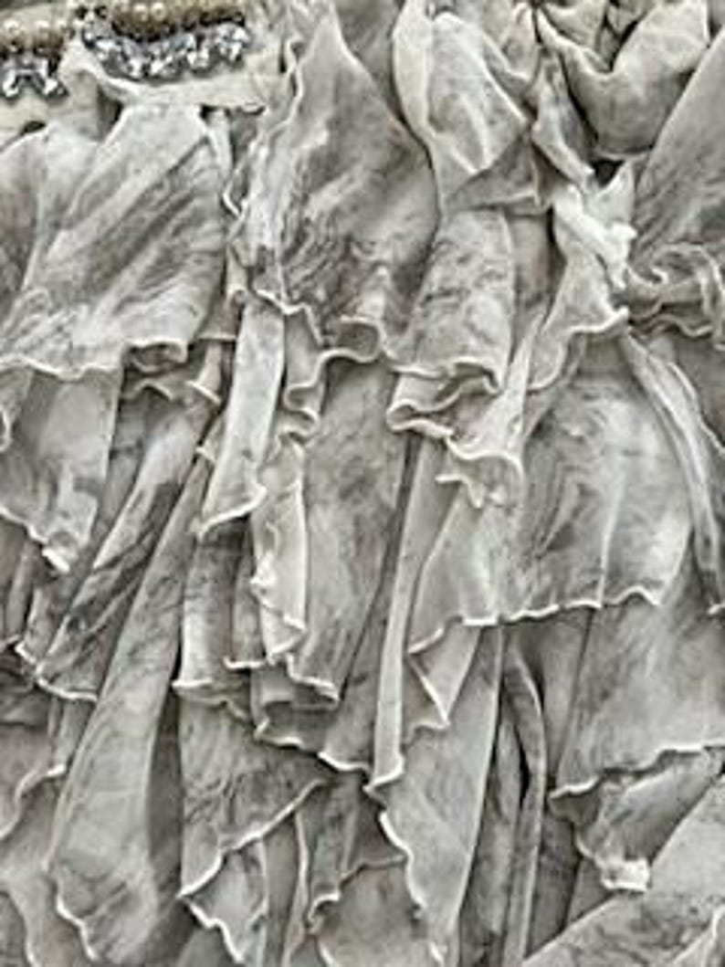 May include: Close-up of a layered, flowing garment with a marbled gray and white pattern. The fabric is sheer and ruffled, creating a textured effect. The top edge features a decorative embellishment with silver and pearl-like beads.