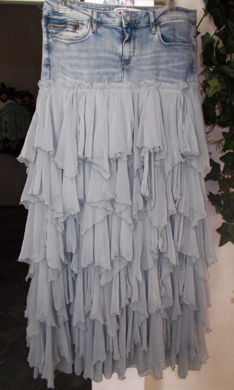 May include: A denim skirt with a light blue wash, featuring a layered, ruffled skirt in a soft, muted blue fabric. The skirt has a classic five-pocket design and a zipper closure.