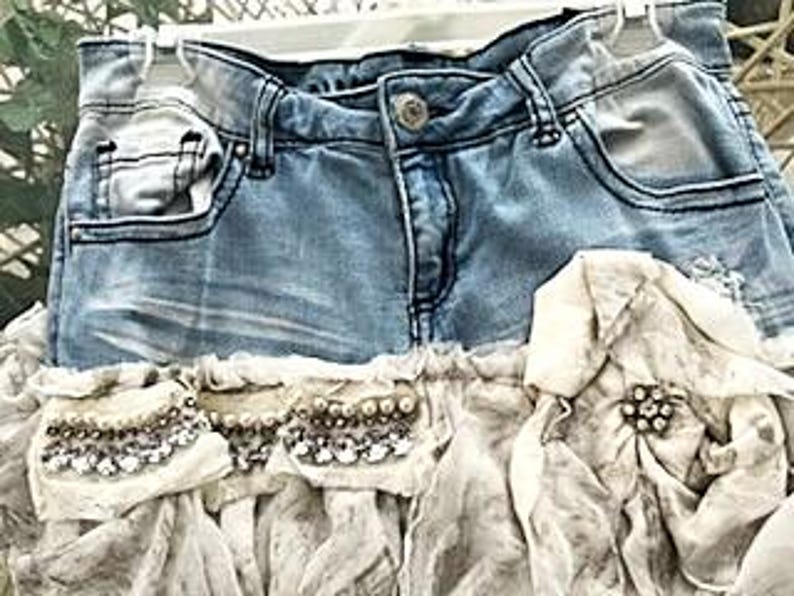 May include: A denim skirt with a light blue wash, featuring a distressed design. The skirt has a ruffled, cream-colored fabric overlay adorned with decorative beading and a central brooch. The skirt is hanging on a white hanger.
