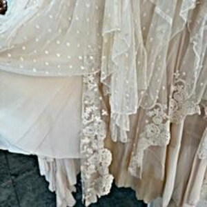 May include: A cream-colored, layered dress with a flowing design. The dress features multiple layers of fabric, including a dotted sheer overlay and lace trim. The dress appears to be made of lightweight materials, with a romantic and ethereal aesthetic.