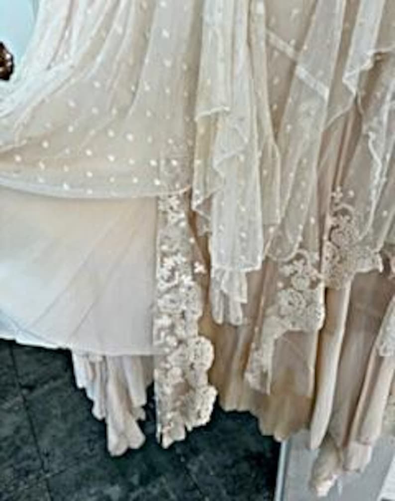 May include: A cream-colored, layered dress with a flowing design. The dress features multiple layers of fabric, including a dotted sheer overlay and lace trim. The dress appears to be made of lightweight materials, with a romantic and ethereal aesthetic.