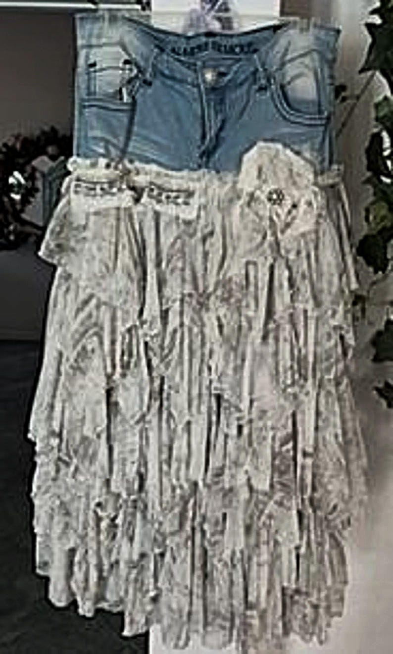 May include: A denim skirt with a light wash, featuring a layered, ruffled design in shades of white and gray. The skirt has a decorative floral accent and a denim waistband with pockets.