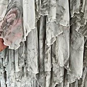 May include: Close-up of a flowing, layered garment with a marbled pattern in shades of gray and white. The fabric appears sheer, with ruffled edges adding texture and movement. The design suggests a dress or skirt.
