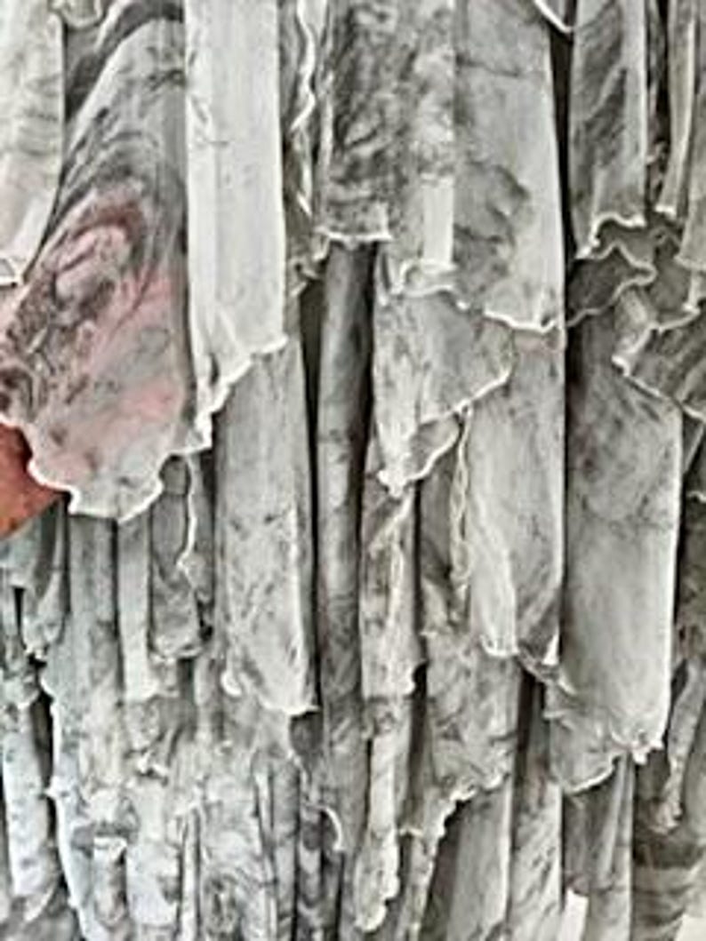 May include: Close-up of a flowing, layered garment with a marbled pattern in shades of gray and white. The fabric appears sheer, with ruffled edges adding texture and movement. The design suggests a dress or skirt.