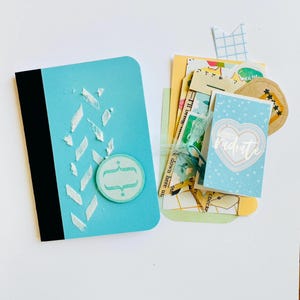 May include: A light blue notebook with a black spine, white textured chevron accents, and a circular tag featuring a bracket symbol. Beside it is a collection of assorted paper ephemera, including a card with a heart and the text radiate.
