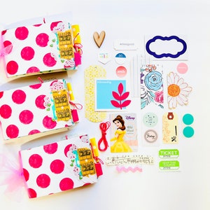 May include: Three pink and white polka dot mini albums with yellow and blue embellishments, a red string, a Disney Belle card, a yellow tag with the text "No. 2", two turquoise circles, a green ticket with the text "Keep this coupon", a white daisy with a yellow center, a blue and white floral pattern, a blue tag with the text "smile", a yellow tag with the text "dreaming", a wooden heart, a blue tag with the text "#feelgood", a white tag with the text "fate", a blue frame, and a pink and white scalloped ribbon.