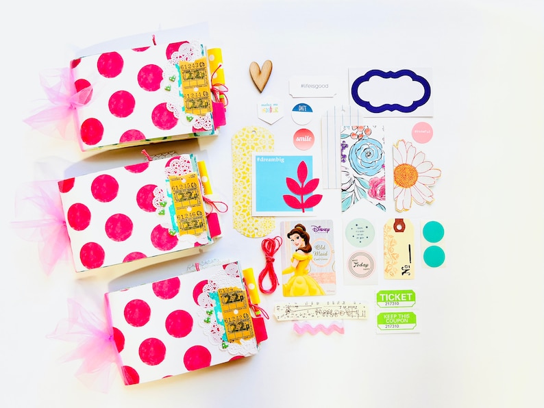 May include: Three pink and white polka dot mini albums with yellow and blue embellishments, a red string, a Disney Belle card, a yellow tag with the text "No. 2", two turquoise circles, a green ticket with the text "Keep this coupon", a white daisy with a yellow center, a blue and white floral pattern, a blue tag with the text "smile", a yellow tag with the text "dreaming", a wooden heart, a blue tag with the text "#feelgood", a white tag with the text "fate", a blue frame, and a pink and white scalloped ribbon.