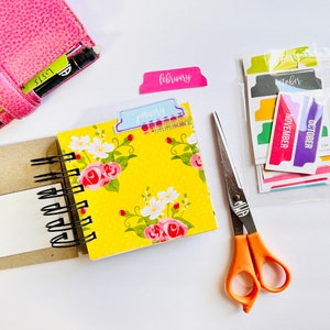 May include: A yellow floral patterned notebook with a black spiral binding. The notebook is open to a blank page. There are colorful month tab dividers in the background, including January, February, October, and November.