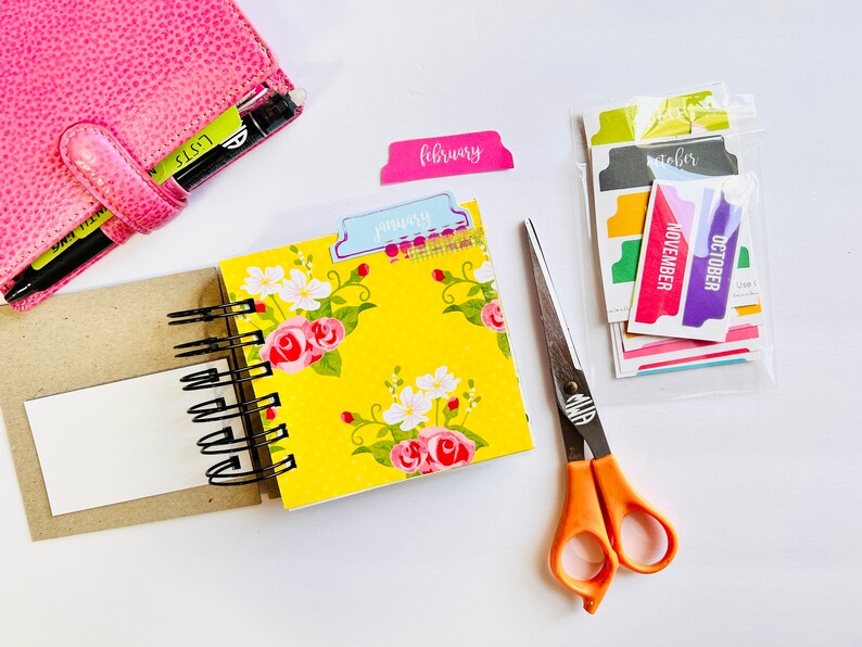 May include: A yellow floral patterned notebook with a black spiral binding. The notebook is open to a blank page. There are colorful month tab dividers in the background, including January, February, October, and November.