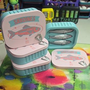 May include: A stack of pink and teal sardine-shaped boxes with a fish illustration and the word "SARDINES". One box is open, revealing three silver fish. The boxes have a pull-tab design. The overall theme is food-related.