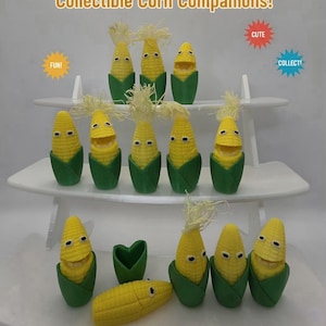 May include: A collection of small yellow corn figurines with green husks and textured kernels, some with open mouths and others with closed mouths, arranged on a white tiered display stand. Text reads MAIZE-IING MATES! Collectible Corn Companions!