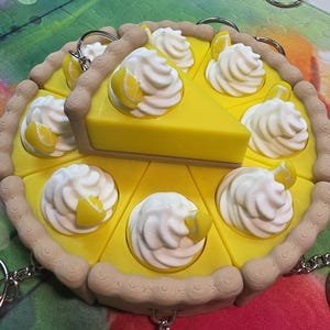 May include: A yellow lemon pie keychain with a slice removed. The pie features white whipped cream dollops with lemon wedges. The crust is tan, and the keychain has a silver chain and ring.