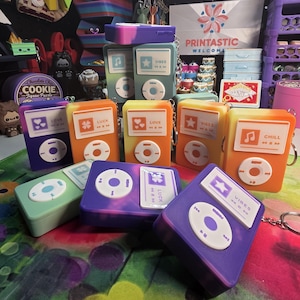 May include: A collection of colorful silicone iPod-shaped keychains. Each keychain features a different design, including musical notes, stars, hearts, and the words "Vibes", "Love", "Luck", and "Chill". The keychains are in shades of purple, orange, yellow, and green.