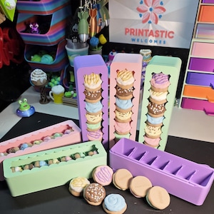 May include: A display of colorful, miniature, decorative cookies and containers. The cookies have various frosting colors and toppings. The containers are rectangular and pastel-colored, with cutouts to hold the cookies. The text "PRINTASTIC WELCOMES" is visible in the background.