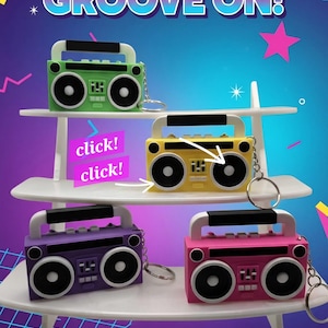 May include: Four boombox keychains in green, yellow, purple, and pink, each featuring a handle, two speakers, and control buttons. The text reads GET YOUR GROOVE ON!, click!, click!, Retro Boombox Keychains, and Available in 4 Cool Colors!