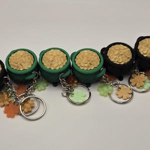 May include: A collection of keychains featuring miniature black and green cauldrons filled with textured, tan-colored material. Each keychain has a silver ring and decorative charms in the shape of flowers and shamrocks.