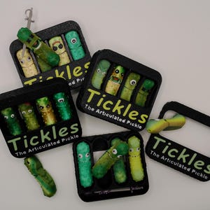 May include: Several black containers with the text "Tickles The Articulated Pickle" are displayed. Each container holds several green and yellow articulated pickle figures with various facial expressions. One pickle is hanging from a silver hook.