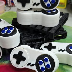 May include: White and black game controller replicas with blue and black buttons. The controllers have a plus-shaped directional pad and two sets of buttons. The controllers are stacked on a black platform.