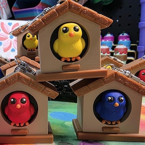 May include: A collection of birdhouse keychains, each featuring a colorful bird inside. The birds are yellow, red, and blue, with black eyes and orange feet. The birdhouses are brown with a small roof and a circular opening.