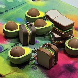 May include: A collection of avocado and sandwich-shaped keychains. The avocado keychains feature a green exterior with a brown pit, while the sandwich keychains have brown bread and colorful fillings. The keychains are on a colorful background.