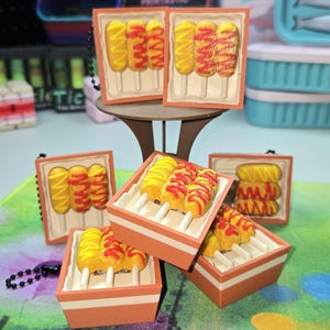 May include: Miniature food charms featuring corn dogs with yellow mustard and red ketchup. The charms are in small orange trays and rectangular frames. Some have keychains attached. The background is colorful.
