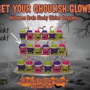 May include: A collection of Halloween brain slushy clicker keychains arranged on a three-tiered white stand. The keychains feature bright neon pink, green, and purple cups with matching brain-shaped tops, straws, and faces. Text reads GET YOUR GHOULISH GLOW! Halloween Brain Slushy Clicker Keyycains, click! click!, and COLLECT ALL 3 SPOOKY COLORS! AVAILABLE NOW!