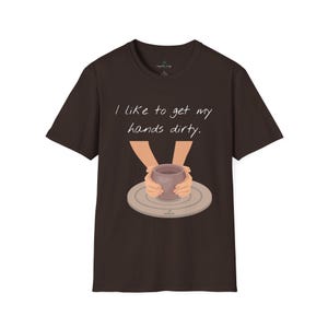 Puede incluir: Una camiseta marrón oscuro con el texto "I like to get my hands dirty". La camiseta presenta un gráfico de manos dando forma a una olla de arcilla en un torno de alfarero. La camiseta es de cuello redondo y manga corta.