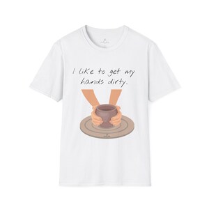 May include: White t-shirt with the text "I like to get my hands dirty." The shirt features an illustration of hands shaping a brown clay pot on a pottery wheel. The design is centered on the front of the shirt.