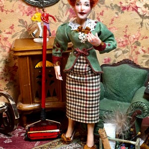 May include: A miniature doll dressed in a green jacket with a white collar and a checkered skirt. The doll has red hair and is standing in a room with a red carpet and floral wallpaper. A red and black carpet sweeper is in front of the doll.
