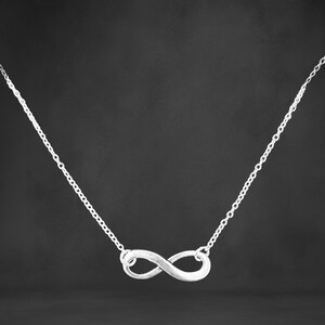 May include: A silver-colored necklace featuring an infinity symbol pendant. The pendant is a stylized figure eight, suspended from a delicate chain. The necklace is displayed against a dark background.