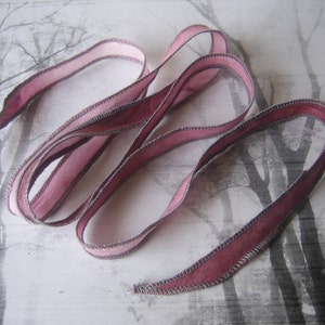 May include: A length of mauve ribbon with a stitched edge, lying in loose loops. The ribbon has a lighter pink hue on one side and a darker mauve on the other. The background features a grayscale image of bare trees.