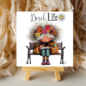 May include: A square print on a small wooden easel features an illustration of a person sitting on a bench, writing. The person has gray hair and a floral crown. The words "Bench Life" are above the illustration.