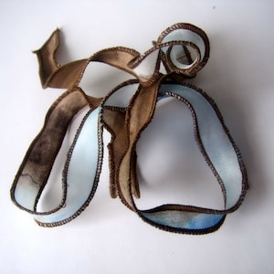 May include: An abstract textile art piece featuring intertwined ribbons. The ribbons are primarily brown and light blue, with dark brown stitching along the edges. The piece has a flowing, organic shape, suggesting movement and texture.