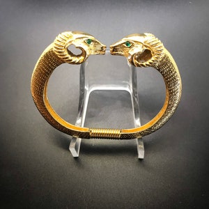 Puede incluir: Un brazalete dorado con bisagras con detalles de cabeza de carnero y ojos de piedras preciosas verdes.