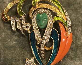 May include: A gold-toned Virgo zodiac sign brooch with a green face, teal and orange accents, and sparkling rhinestones. The brooch features a star and a flowing design, perfect for adding a touch of celestial charm to any outfit.