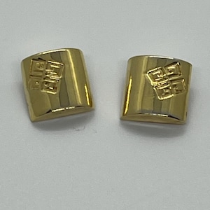May include: A pair of gold-toned square earrings. Each earring features a raised, geometric design in the center. The earrings have a polished, reflective surface, and are set against a neutral background. These earrings are a classic jewelry accessory.