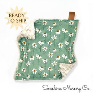 May include: A square, green baby blanket with a daisy pattern. The blanket has a soft, plush, cream-colored border. A brown leather tab is attached to the top corner. The words "READY TO SHIP" are in a sunburst graphic.