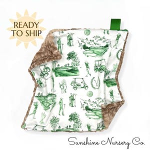 May include: A baby blanket with a white background and a green golf-themed pattern. The blanket has a brown, plush border. The words "Ready to Ship" are in a yellow sunburst shape. The words "Sunshine Nursery Co." are at the bottom.