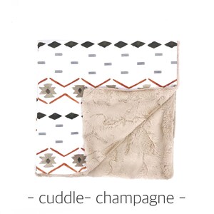 May include: A white and beige blanket with a geometric pattern on one side and a soft, plush texture on the other side. The blanket is folded in half and the pattern side is facing up. The text "cuddle - champagne -" is printed below the blanket.