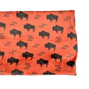 May include: Folded orange fabric with a repeating pattern of black bison silhouettes and arrow-like designs. The fabric has a slightly textured appearance and is folded over, showing the pattern on the top surface.