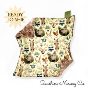 May include: A baby blanket with a woodland animal pattern, including bears, deer, raccoons, and foxes, in shades of brown, green, and cream. The blanket has a soft, plush brown border and a green tag. The text "READY TO SHIP" is visible.
