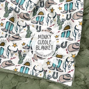 May include: A white Minky Cuddle Blanket with a Western-themed pattern. The design includes cowboy boots, hats, cacti, horseshoes, and bull skulls in shades of turquoise, brown, and tan. The blanket has a green, textured backing.