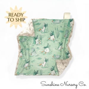 May include: A baby blanket with a duck pond pattern. The blanket features a light green background with illustrations of ducks diving into the water. The border is a soft, plush, tan material. The words "READY TO SHIP" are in a sunburst shape.