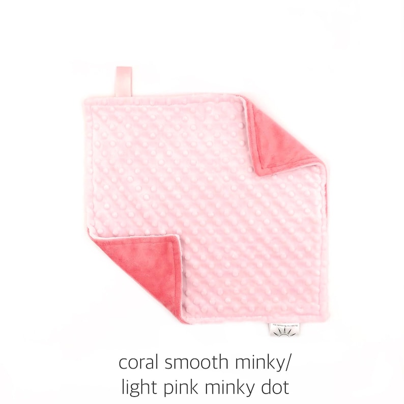 May include: A soft, pink and coral minky baby blanket with a textured dot pattern on one side.  The blanket is made of two different fabrics, one smooth and one with a dot pattern.  The blanket has a loop for hanging.