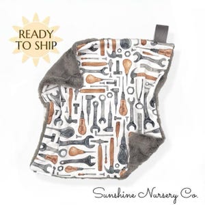 May include: A baby blanket with a white background featuring a pattern of tools in shades of gray and brown. The blanket has a gray plush border and a gray fabric loop. The text "Sunshine Nursery Co." is at the bottom.