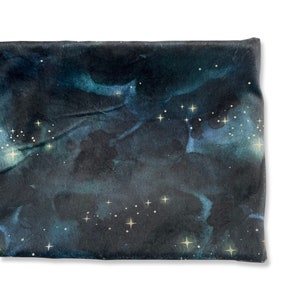 May include: A dark blue fabric with a watercolor galaxy print featuring white and gold stars. The fabric has a soft, textured feel.