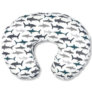May include: A white and gray nursing pillow with a shark print. The pillow is shaped like a crescent moon and is designed to support a baby during breastfeeding or bottle feeding.